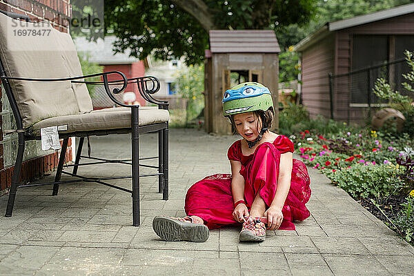 Ein kleines Mädchen in einem schicken roten Kleid und einem Fahrradhelm zieht Sandalen an