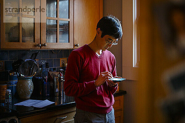 Ein Mann steht in einer Küche bei Fensterlicht und schreibt eine SMS auf einem Mobiltelefon