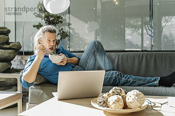 Älterer Mann mit Bart  der auf einem Sofa einen Salat isst und auf seinen Laptop schaut. Pause Zeit Konzept