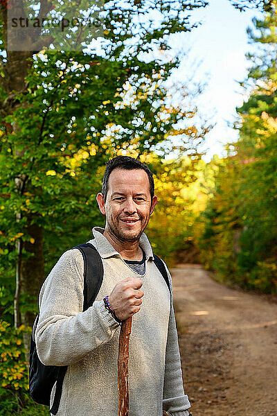 Lächelnder Wanderer  der einen Stock in den Wald hält und in die Kamera schaut