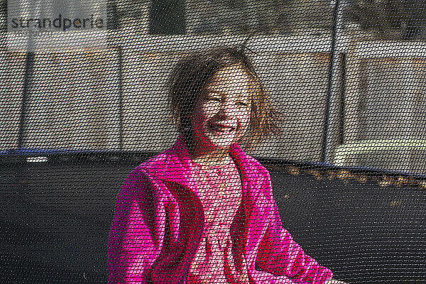 Ein lächelndes kleines Mädchen mit statischem Haar sitzt auf einem Trampolin mit Netz