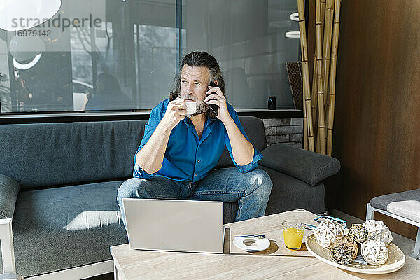 Älterer Mann mit Bart telefoniert und trinkt Kaffee  sitzt auf einem Sofa vor seinem Laptop und schaut aus dem Fenster. Geschäftskonzept