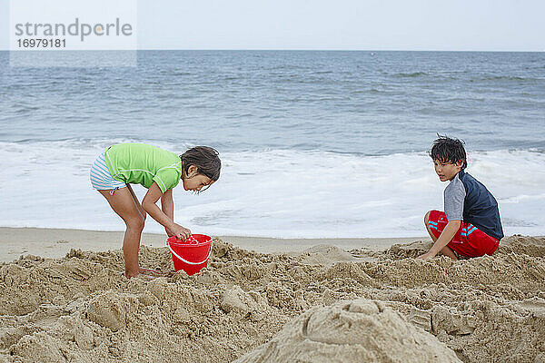 Zwei Kinder spielen zusammen mit einem roten Eimer am Sandstrand am Meer