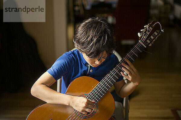 Ein Junge beugt sich über eine klassische Gitarre und spielt einen Akkord im Fensterlicht
