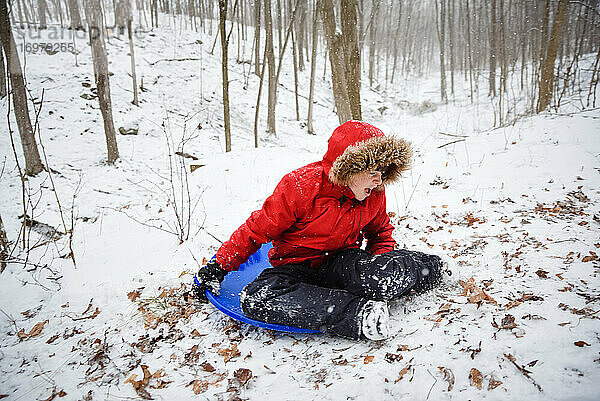 Fröhlicher Junge  der an einem verschneiten Wintertag einen Hügel im Wald hinunterschlittert.