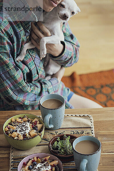 Mensch knuddelt süßen Hundewelpen am Tisch mit gesundem Frühstück und Kaffee