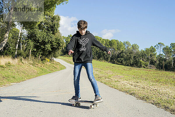 Junger männlicher Teenager mit Kapuze fährt auf einem Skateboard auf einer Bergstraße