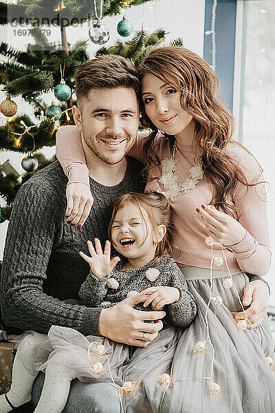 Junge Familie mit einer Tochter in festlicher Kleidung mit einer Girlande in der Nähe des Weihnachtsbaums am Silvesterabend