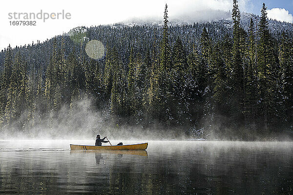 Mann paddelt Kanu auf einem See mit Nebel und Dunst an einem sonnigen Tag im Wald