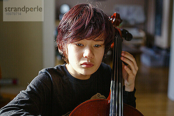 Ein Junge mit intensivem Fokus und rot gefärbtem Haar spielt Cello im Fensterlicht