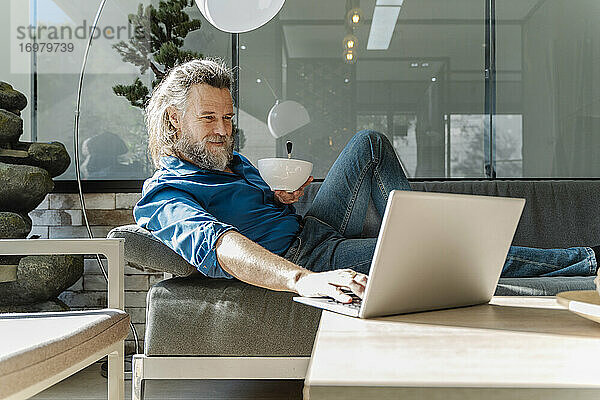 Älterer Mann mit Bart lächelt und arbeitet mit seinem Laptop auf einem Sofa