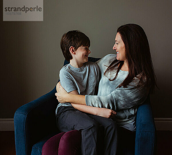 Glückliche Frau  die ihren lächelnden Sohn umarmt  während er in ihrem Schoß auf einem Stuhl sitzt.