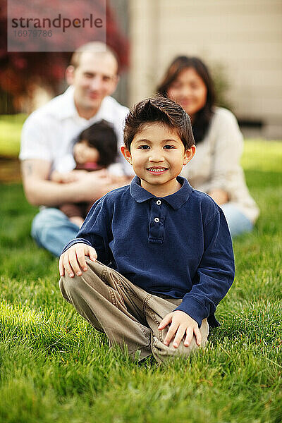 Junge sitzend und lächelnd vor seiner Familie im Gras