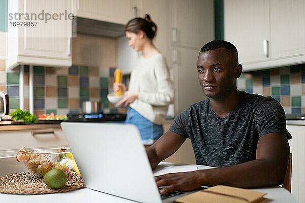 Intelligenter schwarzer Mann arbeitet am Laptop in der Nähe der kochenden Freundin