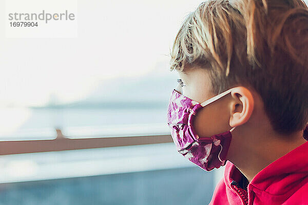 Junge mit einer Maske  der am Flughafen aus dem Fenster schaut.