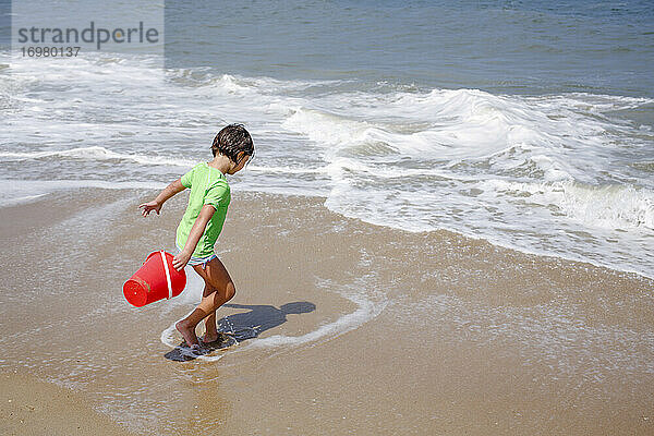Ein kleines Mädchen mit einem Eimer spielt in den Fluten am Rande des Sandstrandes