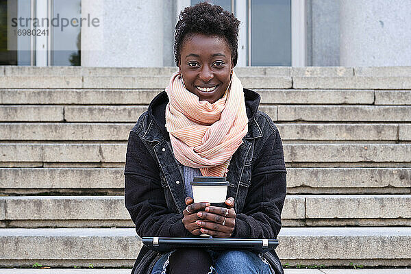 Schöne Universität afrikanische Studentin lächelnd Blick auf die Kamera  hält eine Tasse Kaffee  sitzen auf der Treppe außerhalb auf dem Campus. College Leben Konzept.
