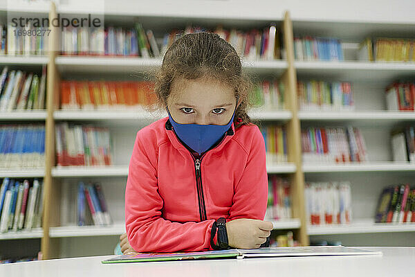 Kleines Mädchen trägt eine medizinische Maske und liest ein Buch in einer Bibliothek. Zurück zu Schule Konzept