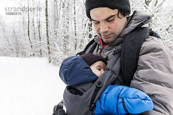 Vater mit süßem Baby in der Trage im verschneiten Winterwald stehend