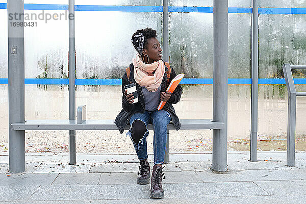 Universität afrikanische Studentin wartet auf einen Bus hält eine Kaffeetasse und einen Ordner auf dem Campus. College Leben Konzept.