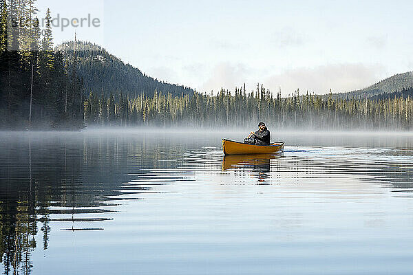 Robuster Mann paddelt Kanu auf nebligem ruhigen See durch dichten Wald
