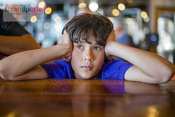 Ein Junge mit dem Kopf in den Händen sitzt am Restauranttisch und wartet geduldig