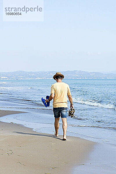 Rückansicht eines Mannes mit Hut und Schuhen  der barfuß am Meeresufer läuft