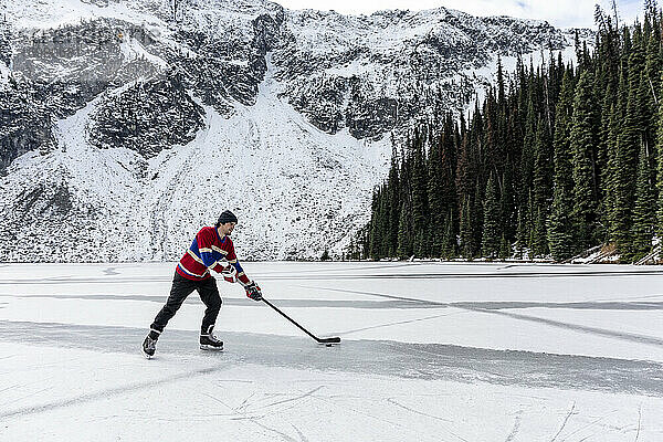Junger Mann spielt Eishockey auf einem See in der Nähe eines schneebedeckten Berges