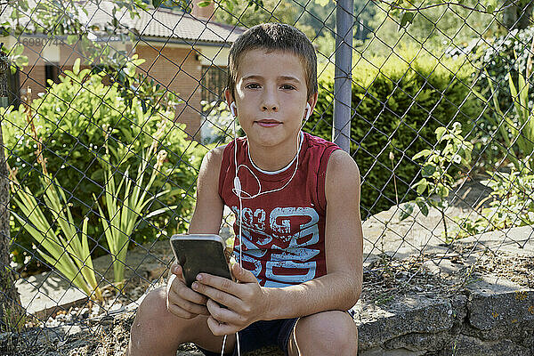 Boy Blick auf ihre Smartphones und Musik hören mit Kopfhörern sitzen auf einer Steinmauer in sonnigen Tag. Technologie-Konzept