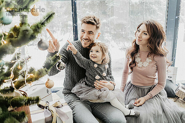 Junge Familie mit einer Tochter in festlicher Kleidung mit einer Girlande in der Nähe des Weihnachtsbaums am Silvesterabend