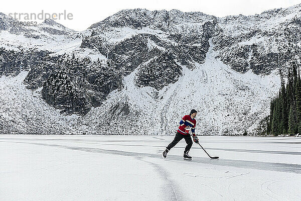 Junger Eishockeyspieler  der auf Schlittschuhen fährt und den Puck mit dem Schläger auf einem zugefrorenen See in der Nähe eines verschneiten Berges und eines Nadelwaldes im Winter in British Columbia  Kanada  manövriert