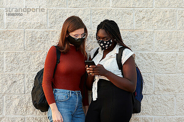 Zwei junge Frauen mit einer Maske schauen auf ihr Smartphone. Multiethnisch