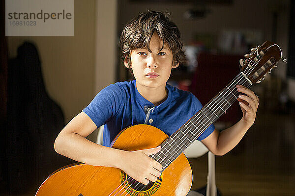 Ein schöner Junge mit intensivem Blick sitzt im Fensterlicht und hält eine Gitarre