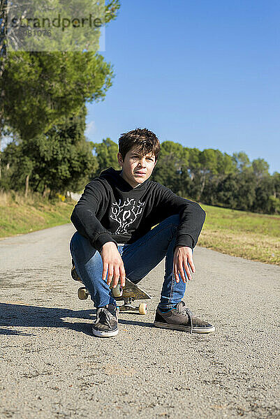 Gutaussehender Teenager mit einem Skateboard auf einer asphaltierten Straße sitzend