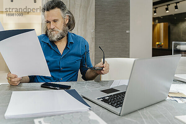 Bärtiger reifer Mann  der an seinem Schreibtisch vor seinem Laptop zu Hause einige Dokumente durchgeht. Business-Konzept
