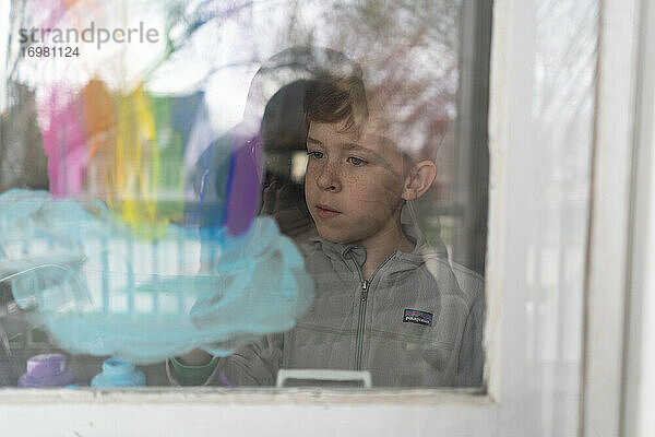 Blick von außen auf einen Jungen  der einen Regenbogen und eine Wolke an ein Fenster malt
