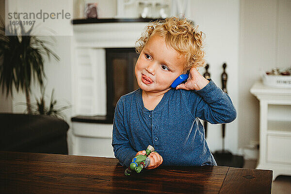 Liebenswerter blonder Junge  der so tut  als würde er mit herausgestreckter Zunge am Spielzeugtelefon sprechen