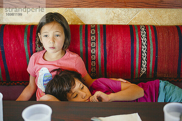 Ein kleines Mädchen sitzt an einem Restauranttisch und hat den Kopf ihres Bruders auf dem Schoß.