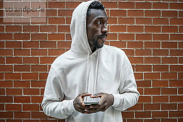 Frontalaufnahme eines afrikanischen Mannes mit ernster Geste und Kapuze  der in der Stadt ein Smartphone benutzt