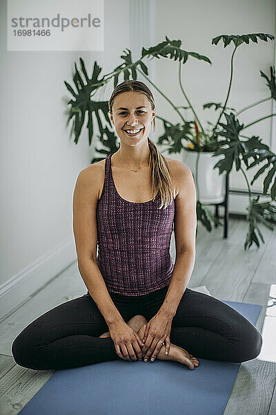 Lächelnder Yogalehrer im Heimstudio  der im Schneidersitz auf einer Yogamatte sitzt