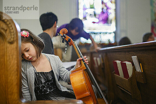 Ein kleines Mädchen in schickem Kleid sitzt in einer Kirchenbank und hält ein Cello