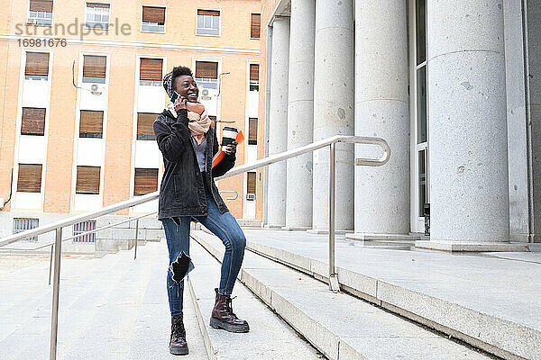 Universität afrikanische Studentin lachend und sprechen auf ihrem Smartphone  hält eine Kaffeetasse auf der Treppe des Schulgebäudes auf dem Campus. College Leben Konzept.