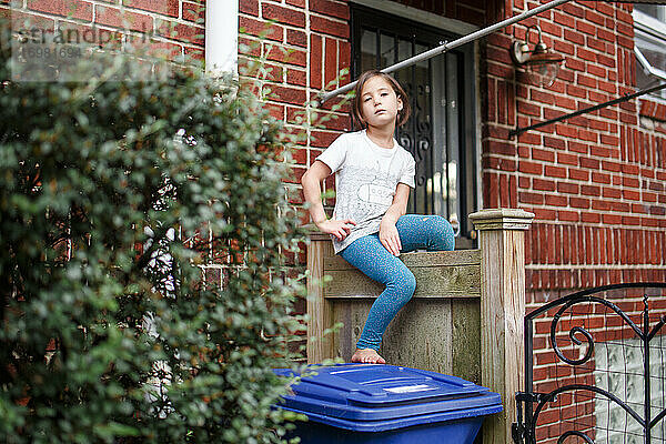 Ein kleines grimmiges Mädchen mit ernstem Blick sitzt auf einem Zaun mit einem Loch in der Hose