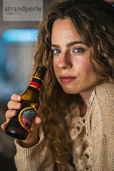 Junge Frau trinkt zu Hause Bier und schaut in die Kamera