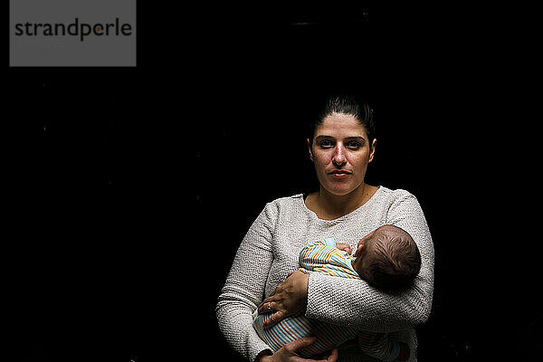 Ruhige Frau  die ein winziges neugeborenes Baby umarmt und in die Kamera schaut  während sie vor einem schwarzen Hintergrund steht