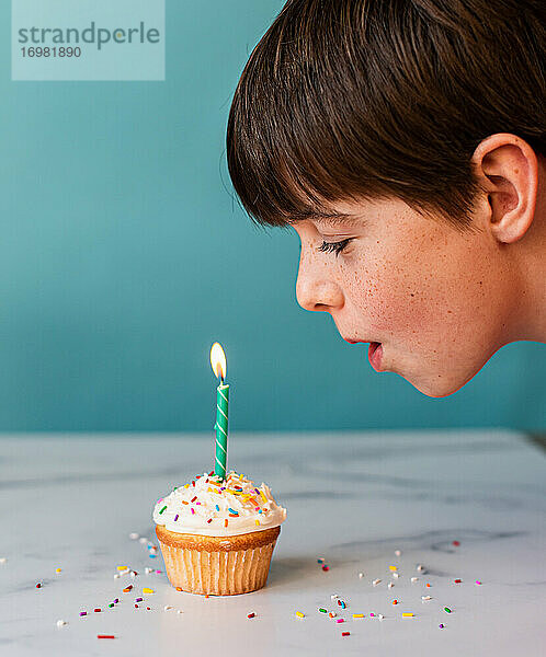 Kleiner Junge bläst eine Kerze auf einem Cupcake mit Zuckerguss und Streuseln aus.
