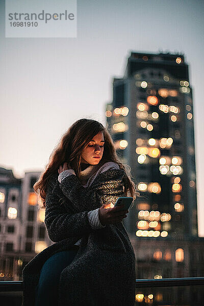 Teenager-Mädchen mit Smartphone sitzt auf Geländer in der Stadt bei Nacht