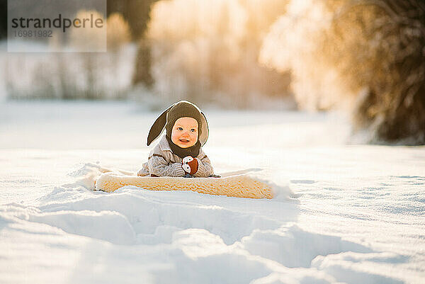 Nettes Baby lächelnd sitzt auf Schnee während Sonnenuntergang in norwegischen Wald