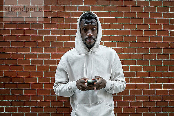 Frontalaufnahme eines afrikanischen Mannes mit ernster Geste und Kapuze  der in der Stadt ein Smartphone benutzt