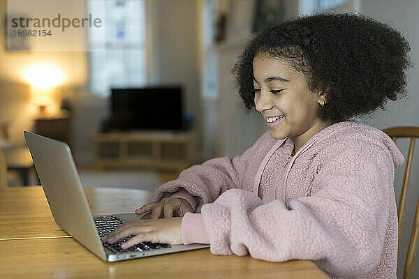 Lächelndes zehnjähriges gemischtrassiges Mädchen arbeitet am Laptop am Tisch